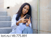 A young woman sits comfortably on stone steps, deeply engrossed in her smartphone, dressed in a casual yet stylish outfit, showcasing the modern lifestyle and its profound connection to technology. A young woman is focused on her smartphone while sitting on steps, enjoying her leisure. Стоковое фото, агентство Ingram Publishing / Фотобанк Лори