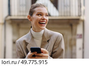 A cheerful young woman happily smiles as she engages with her smartphone, showcasing a joyful and vibrant moment within a busy urban environment during the day while sitting outdoors. Joyful Young Woman Enthusiastically Engaging with Her Smartphone in a Bustling Urban Setting. Стоковое фото, агентство Ingram Publishing / Фотобанк Лори