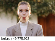A young woman displaying a shocked expression, clad in a stylish blazer over a turtleneck, stands outdoors amidst beautiful greenery, capturing a delightful moment filled with excitement. A surprised young woman joyfully expresses excitement outdoors, surrounded by nature. Стоковое фото, агентство Ingram Publishing / Фотобанк Лори