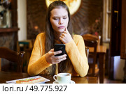 A young woman in a bright orange sweater thoughtfully using her smartphone while enjoying coffee in a caf, surrounded by a warm atmosphere. Young Woman Engaged with Smartphone in Cozy Caf Setting. Стоковое фото, агентство Ingram Publishing / Фотобанк Лори