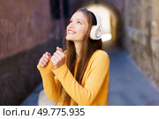 A cheerful young woman with stylish headphones beams with happiness, enjoying her favorite music in a charming alleyway. Her vibrant outfit and joyful expression radiate pure love for music. A Joyful Young Woman Enthusiastically Listening to Music While Strolling in a Scenic Alley. Стоковое фото, агентство Ingram Publishing / Фотобанк Лори