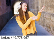 A cheerful and lively young woman, clad in a vibrant yellow sweater joyfully dances with enthusiasm while wearing her favorite headphones in a picturesque urban alley filled with charm. A Joyful Young Woman Delightfully Enjoying Music While Relaxing in an Urban Setting Today. Стоковое фото, агентство Ingram Publishing / Фотобанк Лори