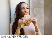 A cheerful young woman with curly hair beams with delight as she takes a satisfying bite of a delicious snack that is wrapped in paper, cherishing her wonderful moment spent outdoors in nature. A woman is enjoying a truly delicious and tasty snack while being outdoors in the fresh air. Стоковое фото, агентство Ingram Publishing / Фотобанк Лори