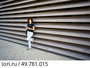 A stylish, young woman with beautiful, curly hair leans casually against a slatted wall, confidently holding a tablet that showcases her trendy urban fashion and her effortless, casual confidence. A casual young woman is leaning against a modern architectural wall, radiating confidence and style. Стоковое фото, агентство Ingram Publishing / Фотобанк Лори