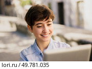 A cheerful young woman sporting a short hairstyle beams with happiness as she actively engages with her laptop in a vibrant, sunlit outdoor environment, relishing every moment of her work. A Young Woman Brightly Smiling While Working on a Laptop in a Beautiful Outdoor Setting. Стоковое фото, агентство Ingram Publishing / Фотобанк Лори