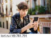 In a vibrant and lively urban area, a young girl dressed in a stylish leather jacket is sitting outdoors, fully absorbed in her smartphone, surrounded by the bustling energy of the city itself. A teenage girl absorbed in her mobile phone in a vibrant urban setting, candidly captured. Стоковое фото, агентство Ingram Publishing / Фотобанк Лори