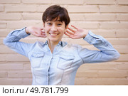 A joyful, vibrant young woman with short hair smiles and poses playfully against a light brick wall, radiating warmth, positivity, and confidence with her cheerful demeanor and carefree attitude. A Cheerful Young Woman with Short Hair Posing in Front of a Stylish Brick Wall Backdrop. Стоковое фото, агентство Ingram Publishing / Фотобанк Лори