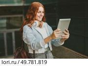 A cheerful and lively teenage girl with vibrant red hair is using a tablet to engage in a fun video call while wearing stylish headphones in a bustling urban environment, surrounded by city life. A Teen Girl Happily Enjoying a Video Call on Her Tablet While in an Urban Outdoor Setting. Стоковое фото, агентство Ingram Publishing / Фотобанк Лори