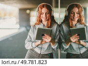 In a lively urban setting, a cheerful young woman with curly red hair finds joy through music, engaging with her tablet and wearing stylish headphones, fully immersed in her entertainment. A Joyful Young Woman is Enjoying Music while Listening on her Tablet with Headphones. Стоковое фото, агентство Ingram Publishing / Фотобанк Лори