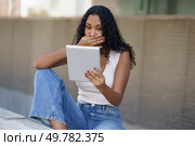 In a lively outdoor scene, a young woman shows surprise, hands covering her mouth in disbelief as she gazes at her tablet, overwhelmed by emotions that capture her attention and curiosity. A Surprised Young Woman Engaging with a Tablet While Sitting in a Beautiful Outdoor Setting. Стоковое фото, агентство Ingram Publishing / Фотобанк Лори