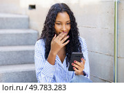 A young woman sits on concrete steps in an urban area, attentively looking at her smartphone with a smile that shows happiness and thoughtfulness, reflecting her curiosity and engagement. A Young Woman is Intently Engaged with Her Smartphone while Sitting on the Steps. Стоковое фото, агентство Ingram Publishing / Фотобанк Лори