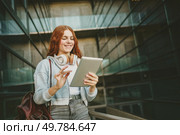 A vibrant young woman with bright red hair confidently holds a sleek tablet and wears trendy headphones, embodying a modern lifestyle in a bustling urban environment with ease and enthusiasm. A young woman is actively engaged with her tablet in a vibrant and modern urban setting. Стоковое фото, агентство Ingram Publishing / Фотобанк Лори