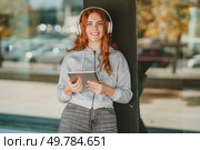 A cheerful young woman featuring curly red hair and trendy headphones beams radiantly with happiness as she holds a sleek tablet, thoroughly immersed in her music in a lively, vibrant outdoor setting. A Young Woman is Enjoying Listening to Music with Headphones While Outdoors in Nature. Стоковое фото, агентство Ingram Publishing / Фотобанк Лори