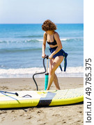 Slim young female redhead in swimwear standing near paddleboard and pumping air with handpump while looking down against waving sea and sky. Female surfer near paddleboard and pumping air. Стоковое фото, агентство Ingram Publishing / Фотобанк Лори