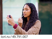 In an urban setting, a fashionable woman with beautiful curly hair elegantly applies her lip gloss while cleverly using her smartphone as a mirror, embodying confidence and chic style. A woman is applying lip gloss while looking at her smartphones mirror, showcasing her beauty. Стоковое фото, агентство Ingram Publishing / Фотобанк Лори