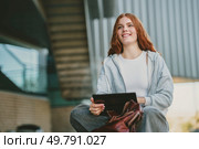In a lively urban environment, a stylish young woman is seated outside, joyfully engaged with her tablet on this stunning sunny day, fully relishing every moment of her vibrant life experience. A Young Woman Delightfully Engaging with Her Tablet in a Vibrant Urban Outdoor Environment. Стоковое фото, агентство Ingram Publishing / Фотобанк Лори