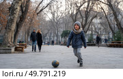 A homeless child playing with a ball in a public park, surrounded by winter trees, while adults walk by without noticing. Стоковое AI-изображение, AI-художник ALLA YURTAYEVA / Фотобанк Лори