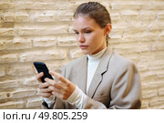 A young woman in a stylish blazer stands confidently against a textured wall, focusing on her smartphone, embodying the essence of modern lifestyle and technology in todays fastpaced world. A focused young woman is fully engaged with her smartphone while in a modern and vibrant setting. Стоковое фото, агентство Ingram Publishing / Фотобанк Лори