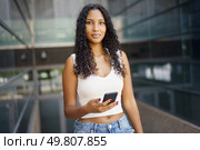 A trendy and stylish young woman dressed in a casual outfit stands confidently in an urban environment. She is holding her smartphone and directing her gaze towards the camera, exuding selfassurance. A Confident Young Woman Holding a Smartphone in a Vibrant Urban Environment, Radiating Energy. Стоковое фото, агентство Ingram Publishing / Фотобанк Лори