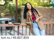 A cheerful young woman with curly hair leans against a railing, holding a phone near a steaming cup of coffee, radiating positivity in a vibrant outdoor setting filled with sunshine. A Joyful Young Woman is Enjoying a Cup of Coffee Outdoors in a Lively Atmosphere and Sunshine. Стоковое фото, агентство Ingram Publishing / Фотобанк Лори