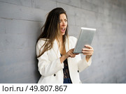 A young woman wearing a stylish white blazer is expressing shock and disbelief while intently looking at her tablet, clearly showcasing a mix of strong emotions against a muted gray background. A shocked woman is reacting to breaking news she is reading on her tablet against a gray wall. Стоковое фото, агентство Ingram Publishing / Фотобанк Лори