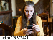 In a cozy and warm cafe setting, a young woman with long flowing hair is sitting comfortably, wearing a bright yellow sweater, completely focused on her smartphone, absorbed in her digital world. A young woman is deeply engrossed in her smartphone while sitting inside a cozy cafe. Стоковое фото, агентство Ingram Publishing / Фотобанк Лори