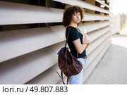 A young woman with beautiful, curly hair is confidently standing against a sleek, modern wall. She holds a phone in one hand and a stylish bag in the other, showcasing a trendy and expressive look. A Young Woman with Beautiful Curly Hair is Leaning Against a Stylish Modern Wall. Стоковое фото, агентство Ingram Publishing / Фотобанк Лори
