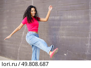 A vibrant young woman dressed in a cheerful pink top and trendy baggy jeans poses joyfully against a textured gray wall, showcasing her playful style, infectious energy, and colorful personality. A Stylish Young Woman Joyfully Dancing in a Colorful Urban Setting, Brimming with Energy. Стоковое фото, агентство Ingram Publishing / Фотобанк Лори
