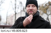 Pensive hipster drinking coffee sitting on a bench in the urban park. Handsome man wearing black coat eating street food, hot dog with paper cup in hand in the city. Stylish bearded middle-aged male. Стоковое видео, видеограф Марина Демидюк / Фотобанк Лори