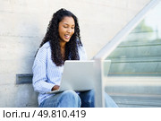 A young woman sits on the stairs, focused on her laptop. Her curly hair and striped shirt represent the productivity and worklife balance many strive for in todays fastpaced world. A Young Woman is Engaged in Working on Her Laptop Outdoors Within a Modern Urban Setting. Стоковое фото, агентство Ingram Publishing / Фотобанк Лори