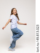 Cheerful genius positive little girl with curly hair in white t-shirt, blue jeans and sneakers has fun and making a step on white background. Happy childhood concept. Copy space. High quality photo. Стоковое фото, фотограф Ирина Михайличенко / Фотобанк Лори