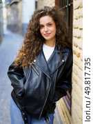 A stylish young woman with curly hair poses effortlessly in a trendy leather jacket, showcasing her keen sense of urban fashion within the picturesque charm of an inviting, vibrant alleyway. A chic young woman in a stylish leather jacket enjoys the vibrant urban atmosphere. Стоковое фото, агентство Ingram Publishing / Фотобанк Лори