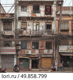 A photograph of an old multi-story apartment building in a densely populated urban area in South Asia (probably India). The building has a dilapidated facade with cracks, peeling plaster, and electrical wires hanging chaotically, forming a dense web over the street. On the lower level are closed shop shutters, small shops, and faded signs. One of the passers-by, an elderly man, sits by a pole, next to a bucket. A scooter is parked in the foreground. The setting conveys the atmosphere of everyday life in the old quarters of the metropolis, emphasizing the contrasts of the urban infrastructure, the fatigue of the buildings, and the saturation of the street environment. old architecture. Стоковое AI-изображение, AI-художник Анна Кривицкая / Фотобанк Лори