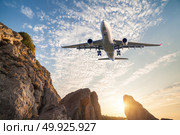Big white aircraft is flying over rocks at sunset. Landscape with passenger airplane, mountains, colorful blue sky with clouds. Passenger airplane is landing. Business travel. Commercial plane. Стоковое фото, агентство Ingram Publishing / Фотобанк Лори