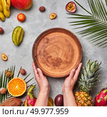 Hands of a woman holding a brown empty plate around a different tropical ripe fruit, coconut, pineapple, physalis, banana on a gray concrete background with copy space. Flat lay. Ripe tropical fruits, coconut, mango, carambola, lychee and palm leaves on a gray concrete background. The woman’s hands hold an empty brown round plate with a copy of the space. Flat lay. Стоковое фото, агентство Ingram Publishing / Фотобанк Лори
