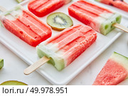 Watermelon ice cream on a stick presented in the form of a square with pieces of kiwi and juicy pieces of watermelon in a plate on a gray marble background. Summer dessert. Close-up of a multicolored fruit frozen smoothies with round pieces of kiwi and triangular pieces of watermelon in a white plate on a gray marble background.Dietary summer dessert. Стоковое фото, агентство Ingram Publishing / Фотобанк Лори