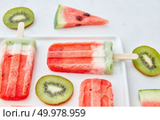 Multicolored fruit frozen smoothies lolly with round pieces of kiwi and triangular pieces of watermelon in a white plate on a gray marble background. Dietary summer dessert. Watermelon ice cream on a stick presented with pieces of kiwi and juicy pieces of watermelon in a plate on a gray marble background. Summer dessert. Стоковое фото, агентство Ingram Publishing / Фотобанк Лори