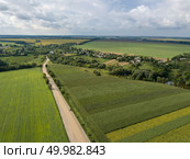 Panoramic view from drone to the countryside with a country buildings, dirt road and agricultural fields against cloudy sky in the summer at sunset.. Aerial view from a bird’s-eye view to a rural landscape with a village, dirt road and agricultural fields of planted crops. Стоковое фото, агентство Ingram Publishing / Фотобанк Лори