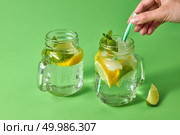 Two glass jars with cold natural cocktail homemade on green background with plastic straw with female hand. Concept of cold alcoholic or non-alcoholic summer drinks.. A woman’s hand inserts plastic straw into a glass jar with lemonade from natural ingredients - water, mint, slices of lemon, lime. Natural cold homemade drinks. Стоковое фото, агентство Ingram Publishing / Фотобанк Лори