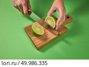 Citrus fruits on a wooden board. Female hands cut with knife a juicy green ripe lime for homemade detox lemonade on a green with copy space.. A woman cut a natural green lime into two pieces on a wooden brown board on a green background. Hands hold a sharp steel knife. Стоковое фото, агентство Ingram Publishing / Фотобанк Лори
