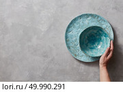 Colorful porceain vintage handmade dishes on a gray marble table with space for text. Woman takes a blue ceramic bowl in her hand . Top view.. Traditional souvenir ceramic handcrafted colorful plate and bowl, holds female hand on a gray concrete table with place under text. Flat lay. Стоковое фото, агентство Ingram Publishing / Фотобанк Лори