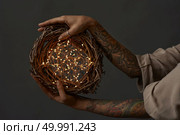 Female hands with tattoos hold a festive natural wreath of branches with a Christmas garland on a dark background. Christmas wreath made of branches decorated with a Christmas lights. Стоковое фото, агентство Ingram Publishing / Фотобанк Лори