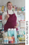 Young pregnant saleswoman wearing burgundy apron standing in personal care and cleaning supplies store aisle, smiling and gesturing warmly, welcoming customers to shopping. Стоковое видео, видеограф Яков Филимонов / Фотобанк Лори