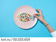 Many different pills and supplements as food on round white plate with woman’s hand. Diet pills and supplements for dieting concept. Flat lay. Colorful capsules and pills on plate in shape of fish with woman’s hand isolated on a blue background and shadows, copy space. Top view. Стоковое фото, агентство Ingram Publishing / Фотобанк Лори