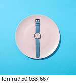 Wristwatch with six o’clock and blue bend on a white plate on a blue background. Time to lose weight, eating control or diet concept. Top view.. Watch with time six o’clock on a white plate on a blue background. Concept of limiting the intake of food diet and weight loss. Flat lay. Стоковое фото, агентство Ingram Publishing / Фотобанк Лори