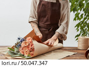 Girl’s florist hands with tatoo are making bouquet with fresh flowers roses living coral color at the table with paper and rope. Process step by step. Concept floral shop.. Florist woman is wrapping fresh rose bouquet in a decorative paper on a wooden table on a light wall background. Process step by step. Mother’s Day holiday. Стоковое фото, агентство Ingram Publishing / Фотобанк Лори