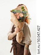 Delicate flowers coral living roses and eryngium as a beautiful bouquet hide girl’s face and ginger hair on a gray background, place for text. Concept of Mother’s or Woman’s Day.. Girl with red hair hide her face big fresh bunch from natural roses and green leaves on a light background. Copy space. Congratulation card. Стоковое фото, агентство Ingram Publishing / Фотобанк Лори
