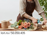 Florist workplace - woman’s hands with tatto are arranging a new bouquet with roses, decorative green leaves on a gray background step by step. Place for text. Concept of flower shop.. Female florist is cutting paper for creating beautiful bouquet on a background of light wall. Place for text. Process of making bouquet step by step. Стоковое фото, агентство Ingram Publishing / Фотобанк Лори