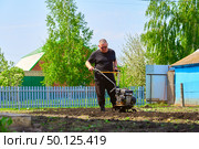 pensioner man with a cultivator cultivates land after winter. Early morning, the sun is just starting to warm the earth, and the walk -behind tractor confidently paves furrows, preparing the ground. Стоковое фото, фотограф Скрипачев Антон / Фотобанк Лори