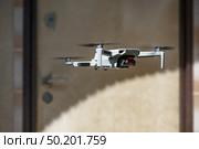 Small Flying Quadcopter Drone in flight mode with camera outdoor in front of the door of a residential building in the shade. Стоковое фото, фотограф Camptoloma / Фотобанк Лори
